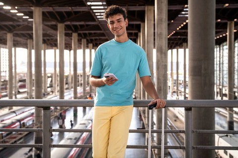 A young traveler in a vibrant blue shirt leans casually against a railing at a train station,