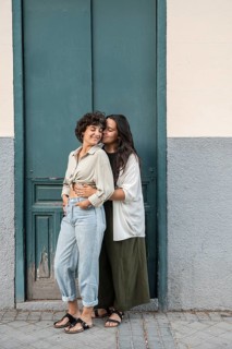 A loving lesbian couple stands in front of a teal door, embracing warmly. One partner gently kisses