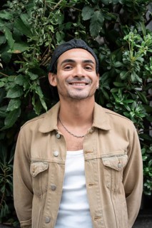 A gay man in casual clothing stands against a lush, leafy background. He sports a chain necklace