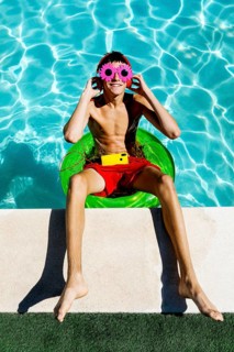 Young teenage boy sits on a green float in a pool, wearing vibrant red shorts, flower-shaped pink