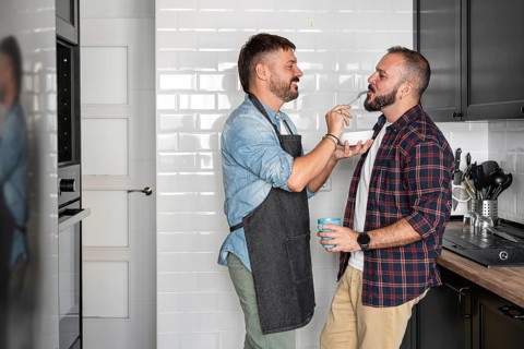 A joyful gay couple shares a moment in the kitchen, one feeding the other with a fork while smiling