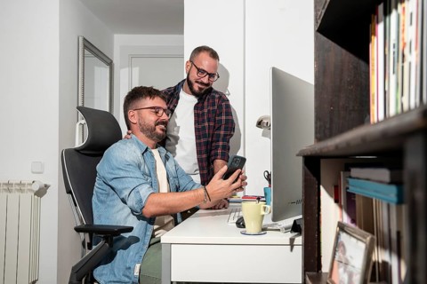 A joyful gay couple shares a moment at their home office. One man sits at a desk, holding a phone,