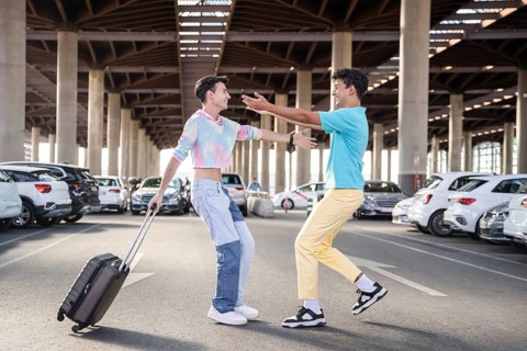 A cheerful gay couple reunites in a spacious parking lot, one carrying a suitcase. They are