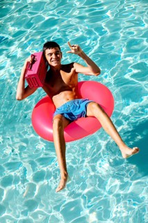 Young teenage boy enjoys a sunny day in a swimming pool, lounging on a pink inflatable ring. He