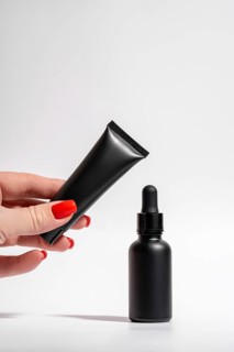 Cropped unrecognizable female hand with red manicure holding a matte black tube and dropper bottle