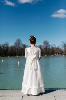 A girl stands by a serene lakeside, wearing a beautiful white First Communion Day dress. Her hair