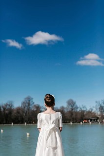 A young girl in a white First Communion dress stands by a tranquil lake, gazing at the serene view.
