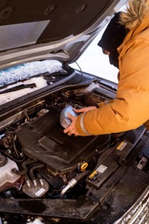 A person in winter clothing checks car fluids in snowy Swedish Lapland. Hood open, engine exposed,