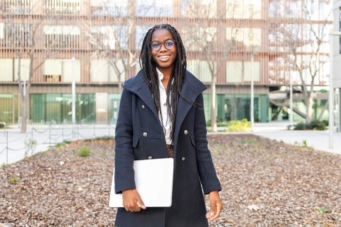 Smiling African American businesswoman, dressed in a stylish coat and glasses, holds a laptop while