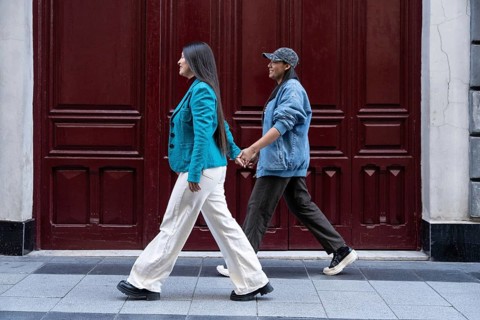 A Latin lesbian couple walks hand in hand along a city street. Their casual attire reflects modern