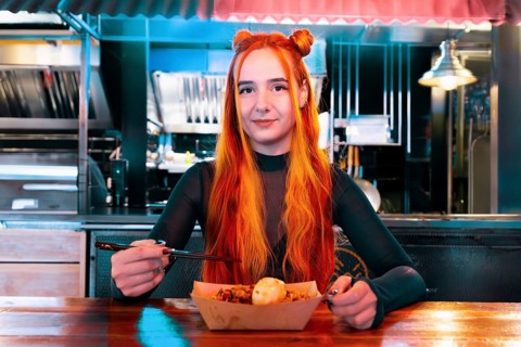 A woman with vibrant orange hair eats using chopsticks at an Asian food market She is seated at a
