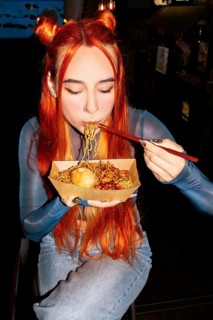 A woman with vibrant hair savoring noodles with chopsticks in an Asian food market, capturing the