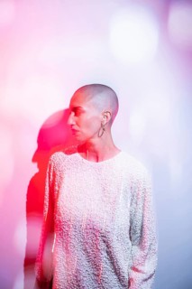 A middle-aged woman with a shaved head, wearing a textured white top, is illuminated by artistic
