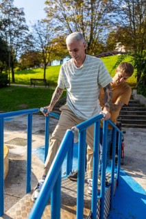 A man with tattoos, holding rails, is assisted by a friend on an accessible ramp in a park Sunlight