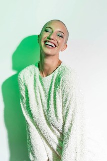 A cheerful middle-aged woman with a shaved head smiles brightly. She wears a textured white