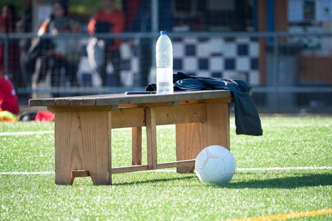 A wooden bench on a soccer field holds a water bottle and jacket, with a ball nearby. Sunlight