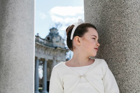A young girl in a beautiful First Communion dress stands gracefully beside a large stone pillar,