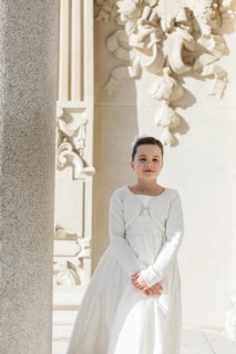A young girl stands in her First Communion dress near an ornate architectural background, embodying