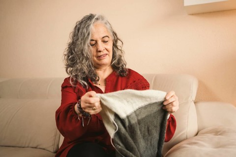 A woman in a red sweater, with gray hair, sits on a beige sofa knitting a gray and white piece. She
