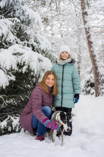 A joyful family poses with a dog in a snowy forest, surrounded by snow-covered trees. The bright