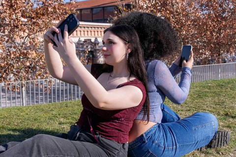 Two young women enjoy a sunny day outdoors while taking selfies. The lady on the left, with fair