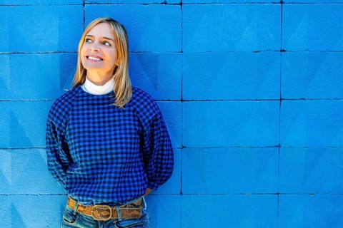 A cheerful woman smiles broadly while dressed in a blue checkered shirt and leaning against a
