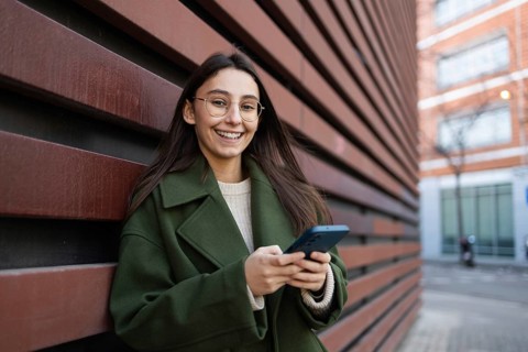 Young woman in a green coat smiling as she uses her smartphone, leaning against a red wall outdoors