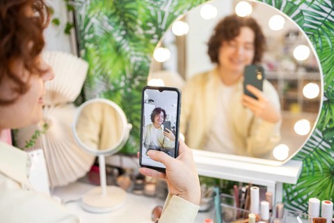 A person takes a selfie using a smartphone in front of a round, illuminated vanity mirror The
