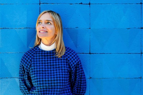 A blonde woman in a plaid shirt gazes upward, standing against a vivid blue background