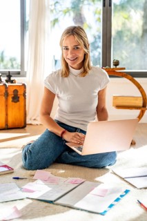 Cheerful woman working from home, sitting cross-legged on the floor with a laptop and paperwork.