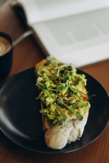 A close-up of avocado toast topped with greens and seeds, served on a black plate. A cup of coffee