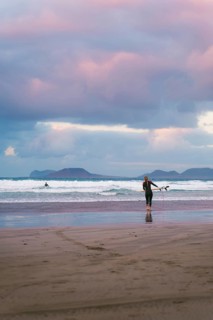 Surfers in a wetsuit approach the waves under a dramatic, colorful sky on a Lanzarote beach. The