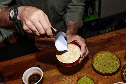 A barista delicately pours steamed milk into a latte, creating intricate latte art. The scene
