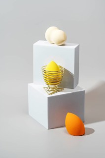 Three cosmetic sponges in different colors rest on minimalist white geometric stands against a soft