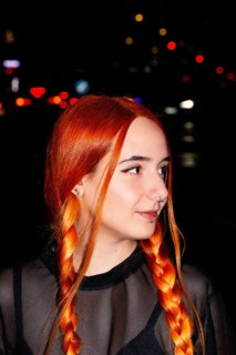 A woman with striking red braids and facial piercings stands against a backdrop of blurred city