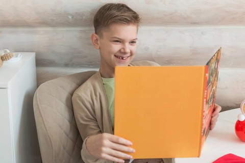 A young boy with short, blond hair enjoys reading a vibrant orange book, his engaging smile