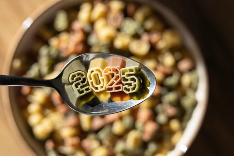 A spoonful of numerically shaped pasta displays the year 2025 above a bowl full of colorful number