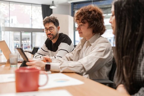 Three coworkers discuss projects around a wooden table in a bright, modern office They share ideas