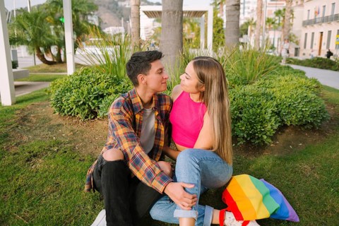 A joyful lesbian couple sits in a park, sharing a loving gaze. With a rainbow flag backpack beside