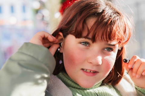 A girl with auburn hair adjusts her earbud while seated on a bus, showcasing a moment of technology