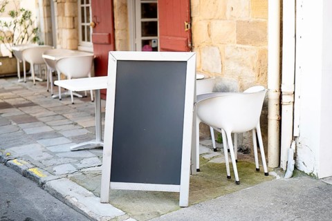A blank chalkboard standing outside a cozy cafe with white chairs and tables, against a stone wall.