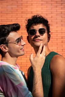 A gay couple shares a tender moment, smiling in the sunlight. They wear sunglasses and casual