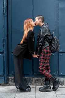 A stylish lesbian couple shares a tender kiss in front of a blue urban background. They exude