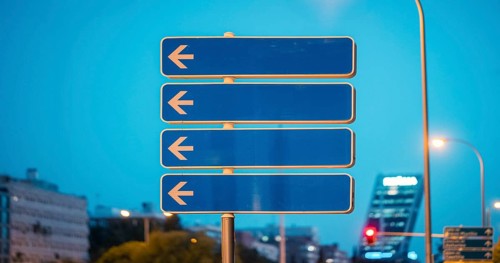 A street sign with multiple arrows pointing to the left, set against a cityscape. The scene