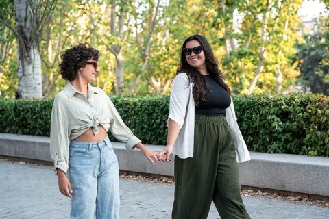 A lesbian couple walks hand-in-hand through a sunny park, enjoying each other's company. The lush