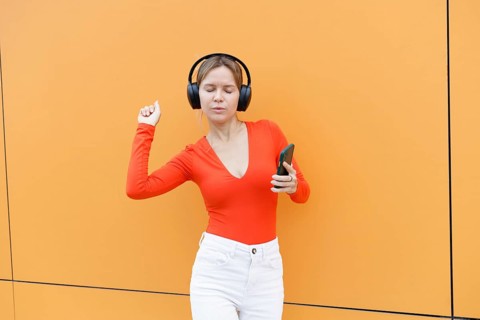 Woman in a red top using a smartphone and wearing headphones, standing against an orange wall in