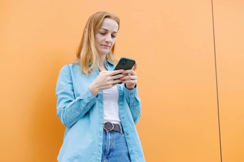 A woman stands against an orange wall in Warsaw, intently using her smartphone She wears a blue