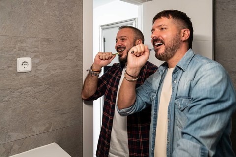 Two gay men in casual shirts joyfully brush their teeth together in a modern bathroom, emphasizing