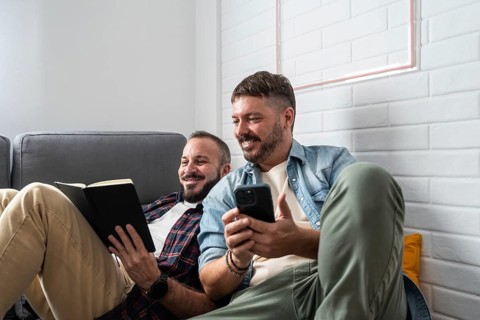 A joyful gay couple relaxes on a cozy sofa, one reading a book and the other holding a smartphone.