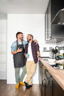 A gay couple standing close in a modern kitchen, sharing a happy moment. One wears an apron and